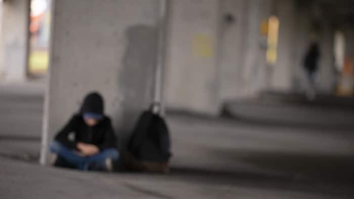 Photo floue d'un adolescent assis en tailleur, capuchon sur la tête et avec un sac à dos, au pied d'un pilier de béton sous un passage supérieur.