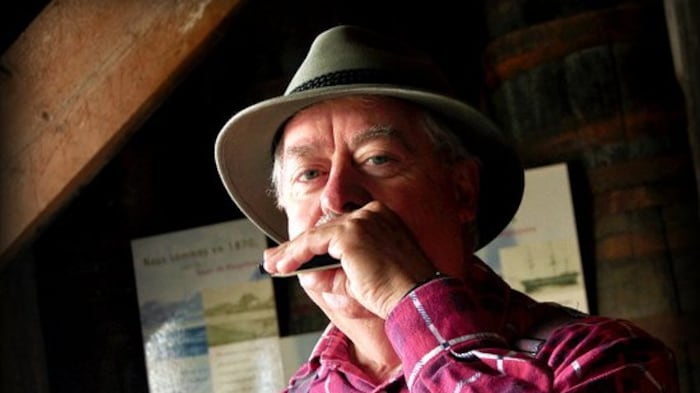 Un homme joue de l'harmonica. Il porte un chapeau beige, une chemise rouge à carreaux et une salopette.