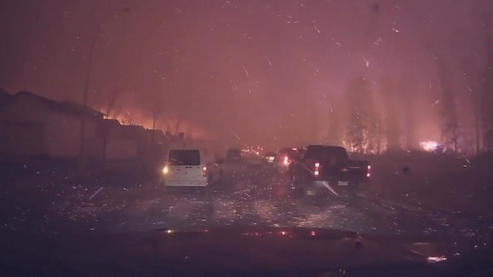 Des voitures qui roulent la nuit sur une route sous une pluie de braises d'un immense incendie tout proche.