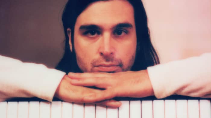 Un homme aux cheveux longs bruns, les mains appuyées sur un clavier dont on voit les notes blanches et noires.