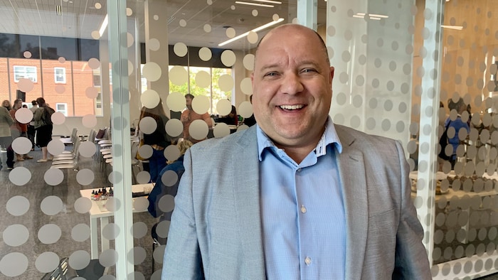 Un homme jovial, vêtu d'une chemise bleue et d'un veston gris, est debout devant un panneau en vitre dans un bureau, et il sourit.