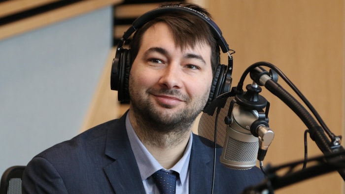 Un homme avec un casque d'écoute sur la tête sourit derrière un micro dans un studio de radio. 
