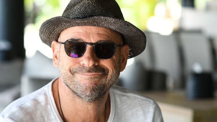 Plan rapproché de Guy Laliberté qui porte des lunettes fumées et un chapeau. Il sourit à la caméra.