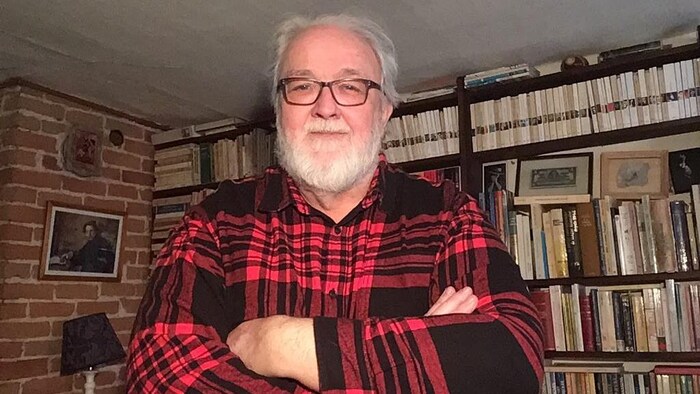 Guy Marchamps se tient devant une bibliothèque remplie de livres, les bras croisés sur la poitrine.