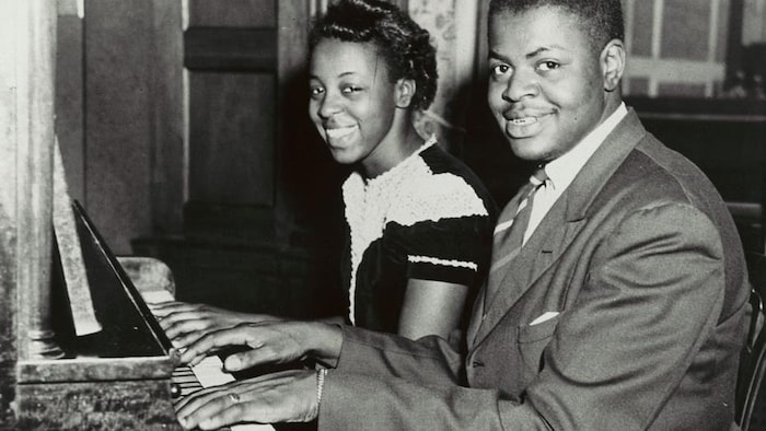 Daisy et Oscar Peterson jouent du piano en souriant à la caméra.