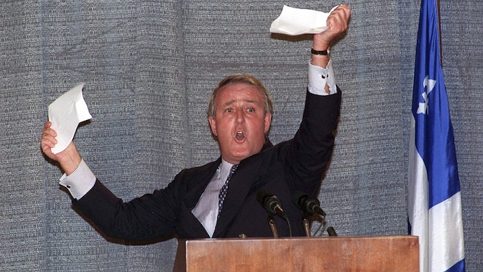 Brian Mulroney tears a paper. A Quebec flag is on his right.