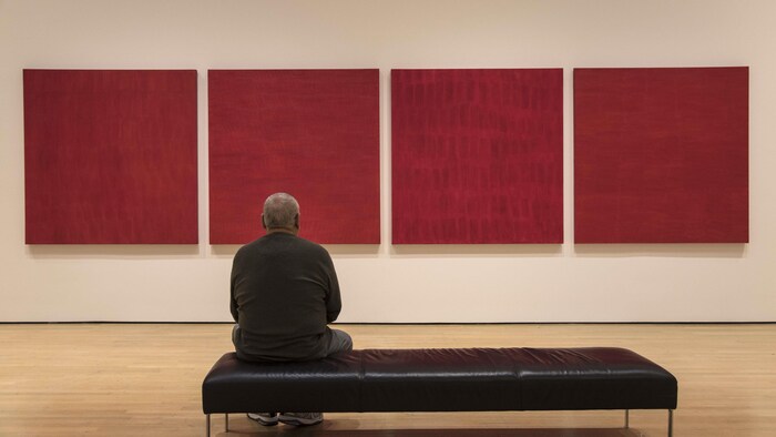 Un homme est assis sur un banc devant l’œuvre composée de 4 tableaux de forme carrée qui sont entièrement rouges.