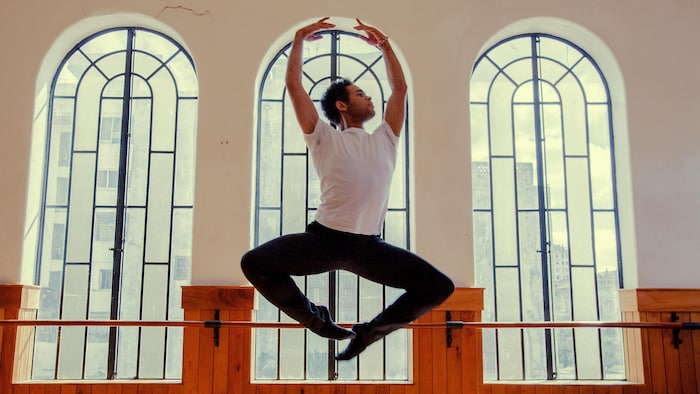 Yonah Acosta Gonzalez en pose de ballet dans les airs devant de grandes fenêtres dans un studio de danse.