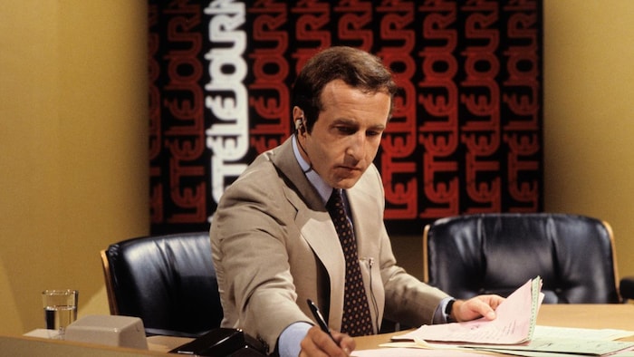 Dans un studio de télévision, l'animateur et journaliste Bernard Derome, assis à un pupitre, prépare son bulletin de nouvelles. À l'arrière-plan, un écran affiche le titre de l'émission à répétition et à la verticale.