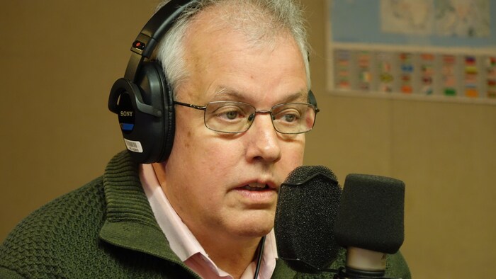 Un homme portant des lunettes et un casque d'écoute est assis dans un studio de radio et parle dans un micro.