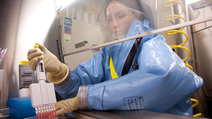 Une jeune femme, couverte d'un scaphandre bleu en caoutchouc qui est gonflé d'air et muni d'une visière transparente, manipule une pipette qui sert à des expériences scientifiques.