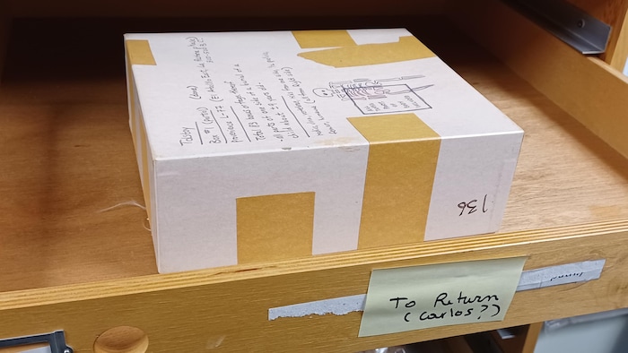 Dans un tiroir de bois, dans un laboratoire d'anthropologie, se trouve une boîte, sur laquelle apparaissent des inscriptions et un dessin de squelette.