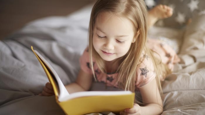 Une petite fille lit un livre, allong&eacute;e sur un lit.