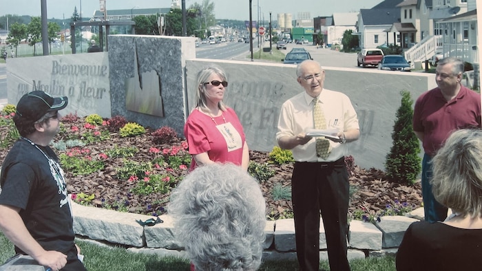 Un public écoute un discours devant une pancarte de pierre où un peu lire « Bienvenue au Moulin à fleur », aux abords d'une rue principale.