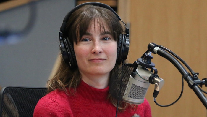 Une femme avec des écouteurs esquisse un léger sourire devant un micro dans un studio de radio. 
