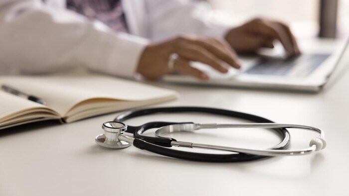 Un stéthoscope au premier avec en fond, un cahier, un ordinateur et les mains d'un médecin qui tapent sur le clavier.