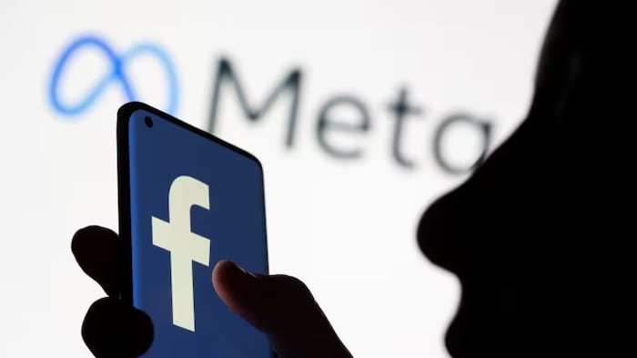 La silhouette d'une personne tenant un cellulaire sur lequel figure le logo de Facebook et celui de Meta en arrière-plan.

