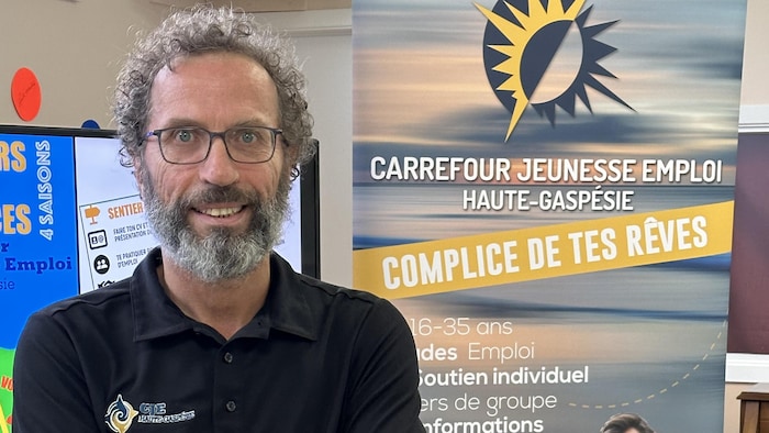 Michel Rioux les bras croisés devant l'affiche de la Journée de l'emploi.