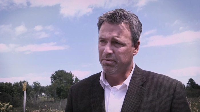 Mike Holland, concentré, écouter une question d'un journaliste, dans un extérieur avec ciel bleu.