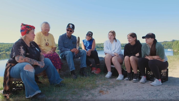 Mique Michelle assise devant la rivière Mattagami, accompagnée de deux aînés et quatre jeunes.