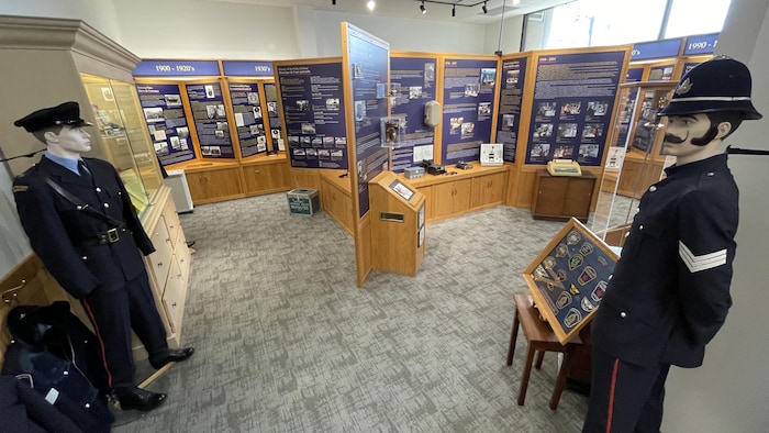 Une section du musée de la police de Sudbury situé au 128 rue Larch. En avant-plan, il y a une mannequin en uniforme et un cadre avec des badges. En arrière-plan, il y a des panneaux avec du texte et des objets divers.