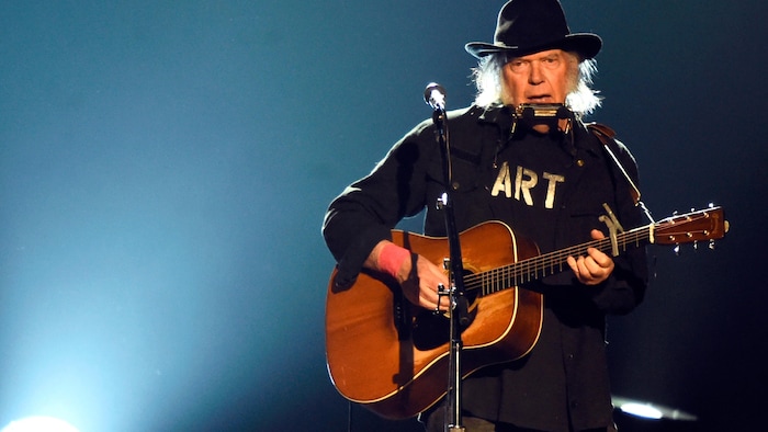 Neil Young joue de la guitare et de l'harmonica derrière un micro, sur scène.