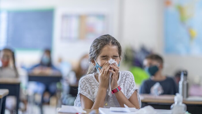 Une élève de l'élémentaire abaisse son masque pour se moucher. Ses pairs sont assis à leur pupitre individuel en arrière-plan.