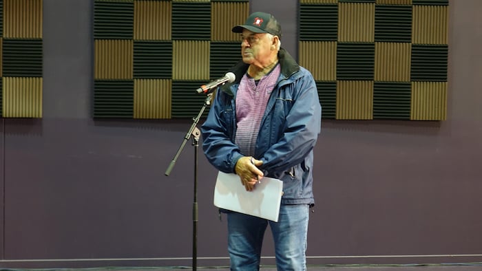 Un homme debout parle au micro portant un manteau et une casquette.