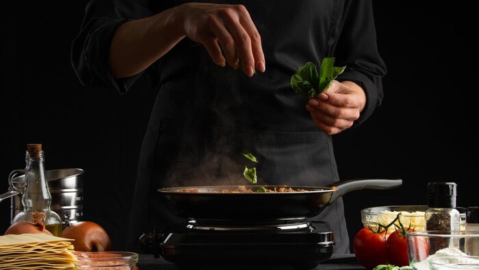 Une chef met du basilic dans une poêle avec un plan de travail garni de tomates, de pâtes, d'oignons et d'huile d'olive.  