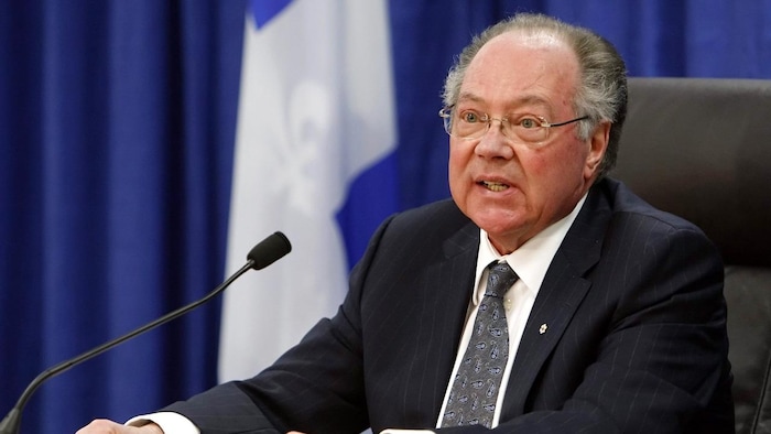 Photo de Michel Bastarache devant un drapeau du Québec, en train de parler dans un micro.