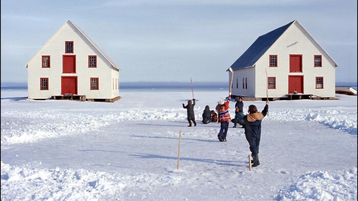 Des personnes jouent au hockey sur la glace enneigée devant deux maisons.