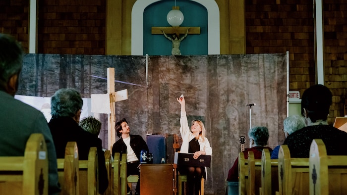 Deux comédiens, un homme et une femme, en train de jouer devant un public dans une église. Derrière eux, un décor qui évoque une forêt.