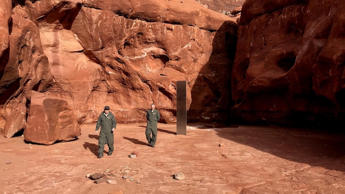 Un monolithe en métal est planté dans le désert et deux hommes marchent aux alentours. 