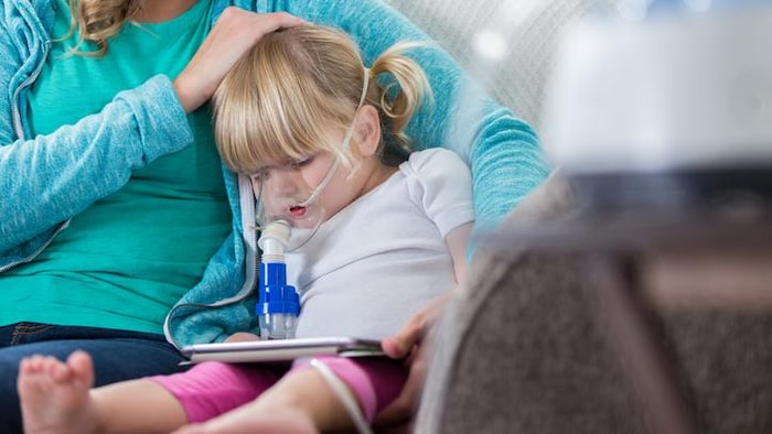 Une petite fille souffrant de fibrose kystique respire dans un masque afin de dégager ses voies respiratoires; elle est assise à côté de sa mère qui lui caresse la tête.  