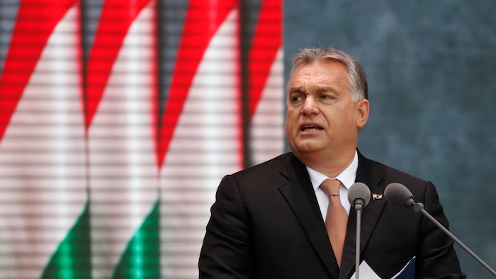 Viktor Orban debout devant un podium, des drapeaux hongrois en arrière plan.