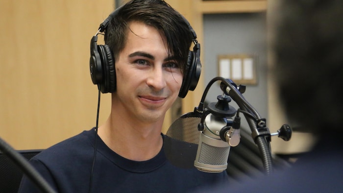Un homme, casque d'écoute sur la tête, regarde en souriant dans la direction de l'animateur dans un studio de radio.