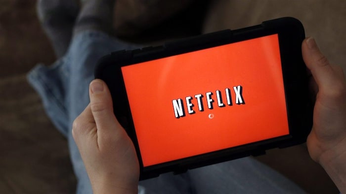 L'écran d'une tablette tenue par un homme, dont on aperçoit les pieds en arrière-plan, affiche le logo de Netflix.