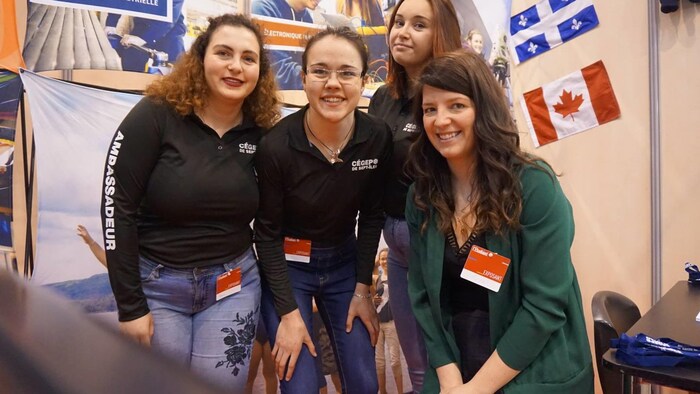 Les quatre femmes sourient à la caméra près d'un kiosque faisant la promotion d'études au Québec. 
