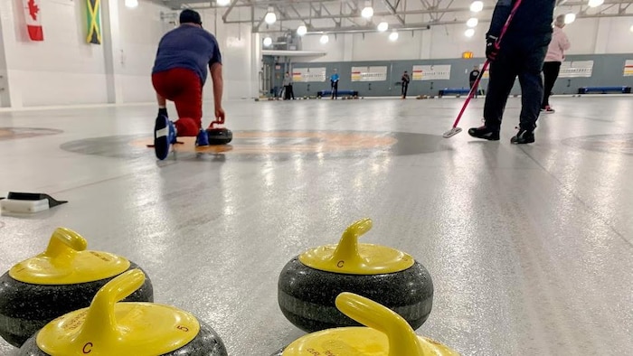 On voit des pierres de curling sur la glace et une personne en arrière-plan qui en lance une.