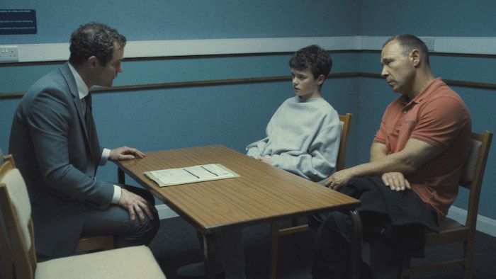 Dans une petite pièce, un homme en veston est assis d'un côté d'une table et un jeune garçon et son père sont assis de l'autre côté, l'air sérieux.