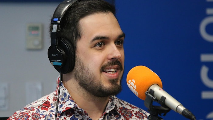 Un homme portant un casque d'écoute et une chemise à motif coloré parle devant un micro dans un studio de radio. 