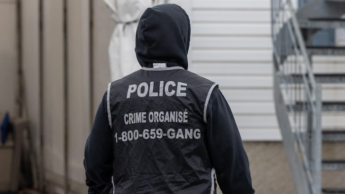 Un policier vêtu d'un dossard où il est indiqué : police, crime organisé avec un numéro de téléphone, est dehors, devant une maison.