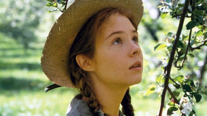 Anne Shirley (Megan Follows) coiffée d'un chapeau de paille se tient près d'un arbre en fleurs.