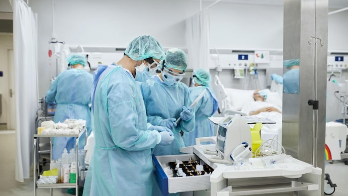 À l'avant-plan, deux professionnels de la santé manipulent des fournitures médicales. Derrière eux, d'autres s'occupent d'un patient dans un lit d'hôpital. Tous portent des vêtements protecteurs.