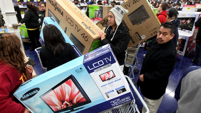 Des clients dans un magasin d'électronique attendent en file à la caisse pour payer. La plupart ont acheté un téléviseur.