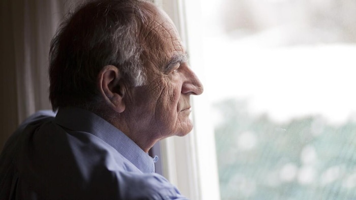 Un homme âgé seul regarde depuis une fenêtre vers l'extérieur.