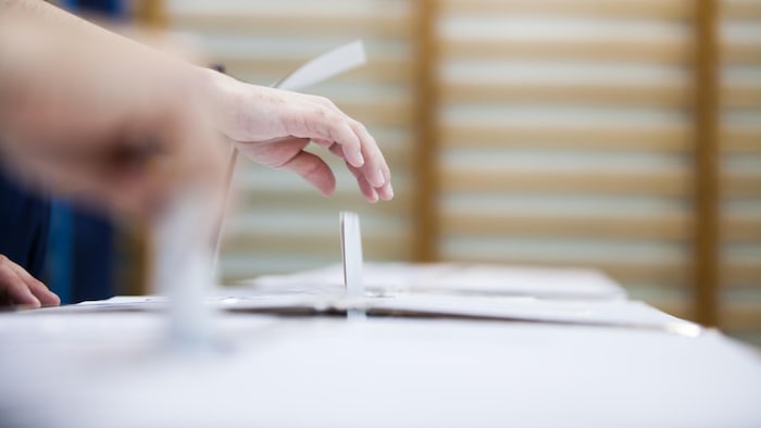Gros plan sur des mains qui déposent un bulletin de vote dans une urne en carton, dans un gymnase.