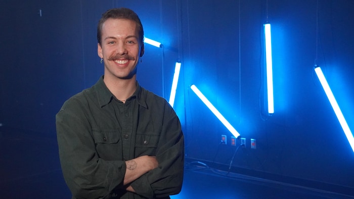 Un homme souriant qui pose sur une scène. Plusieurs lumières bleues sont suspendues derrière lui.
