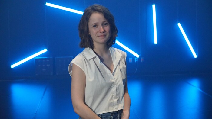 La femme souriante pose sur une scène. Plusieurs lumières bleues sont suspendues derrière elle.