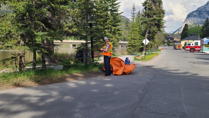 Un employé municipal se tient près d'une grande bâche orange qui n'a pas encore été déployée. Des camions de pompiers peuvent être observés le long de la rivière, à Banff, le 28 mai 2026.
